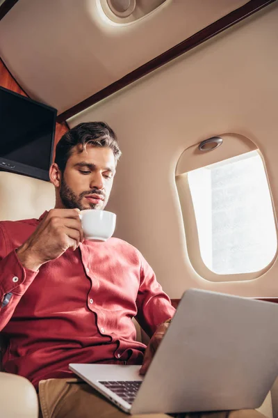Handsome man in shirt using laptop and holding cup in private plane — Stock Photo