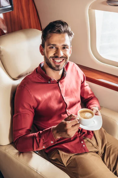 Hombre guapo en camisa sonriendo y sosteniendo taza en avión privado - foto de stock
