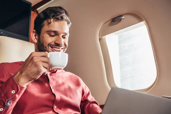 Улыбающийся мужчина в рубашке с помощью ноутбука и держа чашку в частном самолете — стоковое фото