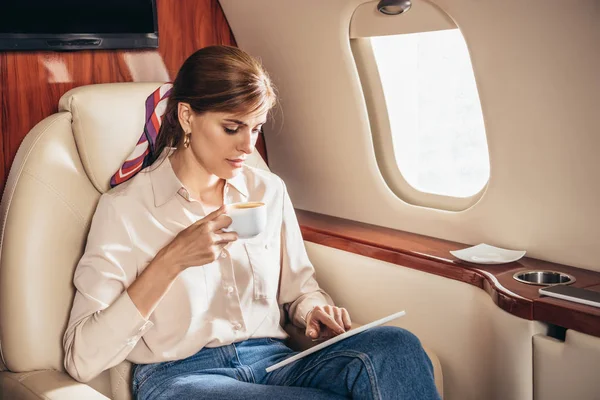 Mulher atraente na camisa usando tablet digital e segurando xícara de café no avião privado — Fotografia de Stock