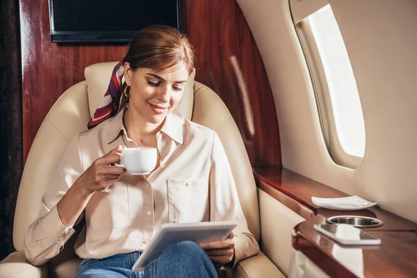 Привлекательная женщина в рубашке с помощью цифрового планшета и проведение чашку кофе в частном самолете — стоковое фото