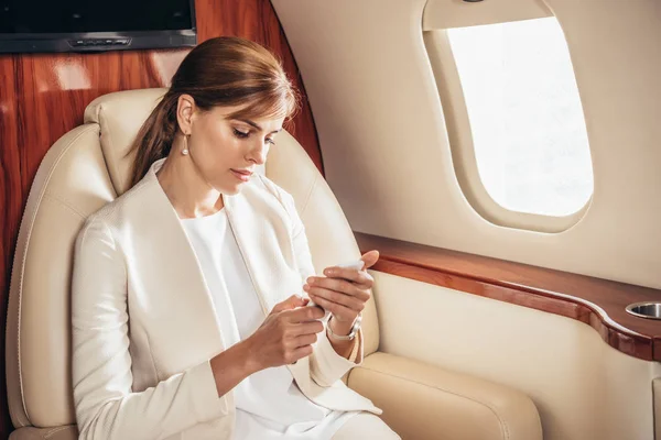 Attractive businesswoman in suit using smartphone in private plane — Stock Photo