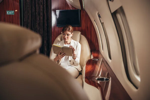 Selective focus of attractive businesswoman in suit reading book in private plane — Stock Photo