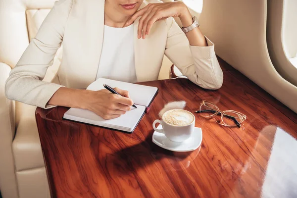 Cropped view of businesswoman in suit writing in notebook in private plane — Stock Photo