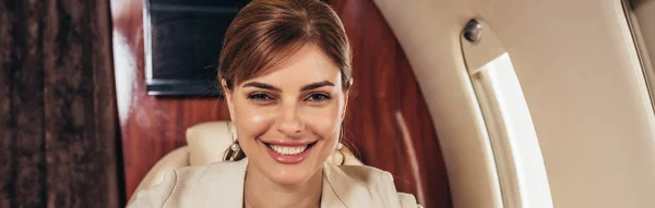 Tiro panorâmico de mulher de negócios atraente sorrindo e olhando para a câmera no avião privado — Fotografia de Stock
