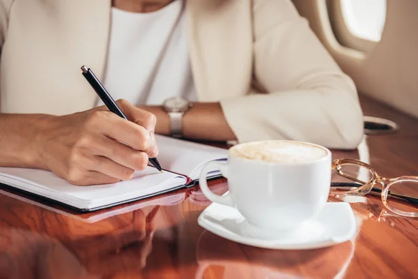 Cropped view of businesswoman in suit writing in notebook in private plane — Stock Photo