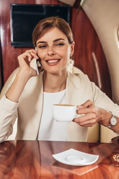 Attractive businesswoman in suit holding cup and talking on smartphone in private plane — Stock Photo