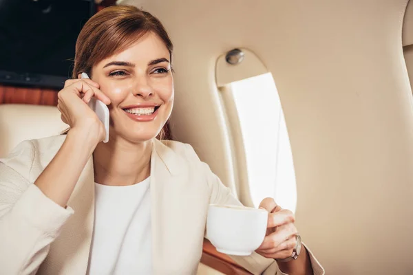 Smiling businesswoman in suit holding cup and talking on smartphone in private plane — Stock Photo