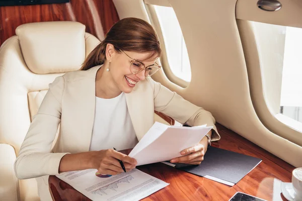 Усміхнена бізнес-леді в костюмі робить паперові роботи в приватному літаку — стокове фото