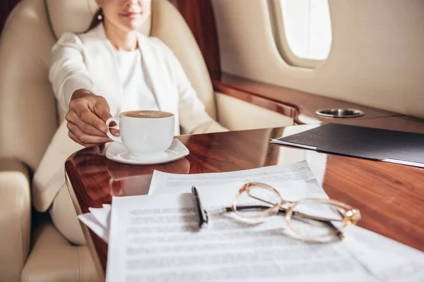 Cropped view of businesswoman in suit taking cup in private plane — Stock Photo