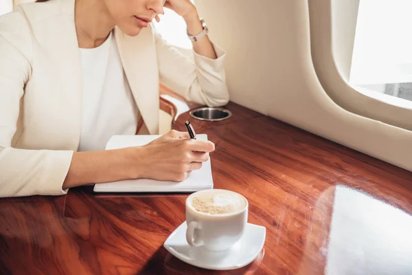 Vista recortada de la mujer de negocios en traje escrito en cuaderno en avión privado - foto de stock