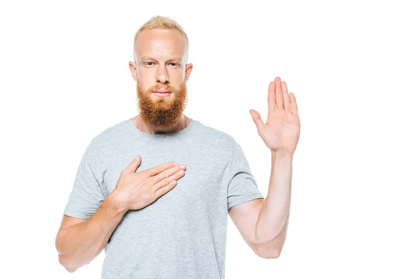 Ritratto di un uomo barbuto serio che dà un giuramento con mano in alto e mano sul cuore, isolato su bianco — Foto stock