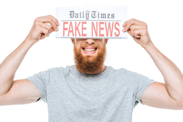 Hombre barbudo sonriente sosteniendo el periódico con noticias falsas delante de los ojos, aislado en blanco - foto de stock