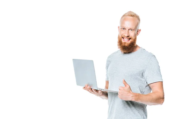 Allegro uomo barbuto utilizzando il computer portatile e mostrando pollice in alto, isolato su bianco — Foto stock