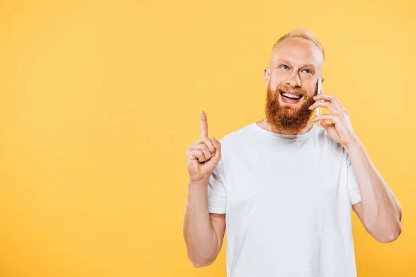 Sonriente hombre barbudo que tiene idea mientras habla en el teléfono inteligente, aislado en amarillo - foto de stock
