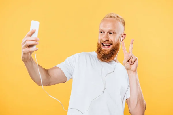 Happy bearded man with earphones taking selfie on smartphone and showing victory sign, isolated on yellow — Stock Photo