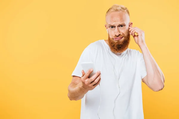 Skeptical bearded man listening music with earphones and smartphone, isolated on yellow — Stock Photo