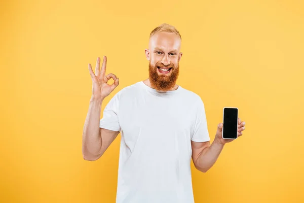 Hombre barbudo sonriente mostrando signo de ok y teléfono inteligente con pantalla en blanco, aislado en amarillo - foto de stock