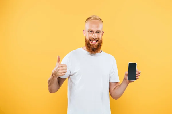 Hombre barbudo mostrando el pulgar hacia arriba y teléfono inteligente con pantalla en blanco, aislado en amarillo - foto de stock