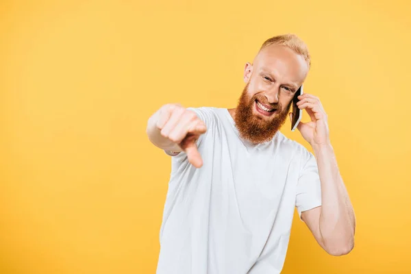 Hombre barbudo alegre hablando en el teléfono inteligente y apuntando a usted, aislado en amarillo - foto de stock