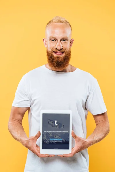 KYIV, UCRANIA - 27 de agosto de 2019: hombre barbudo sonriente mostrando una tableta digital con aplicación tumblr en la pantalla, aislado en amarillo - foto de stock