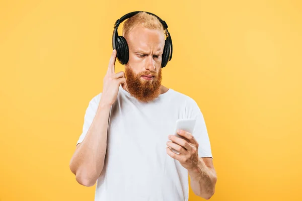 Pensoso uomo barba ascoltare musica con cuffie e smartphone, isolato su giallo — Foto stock