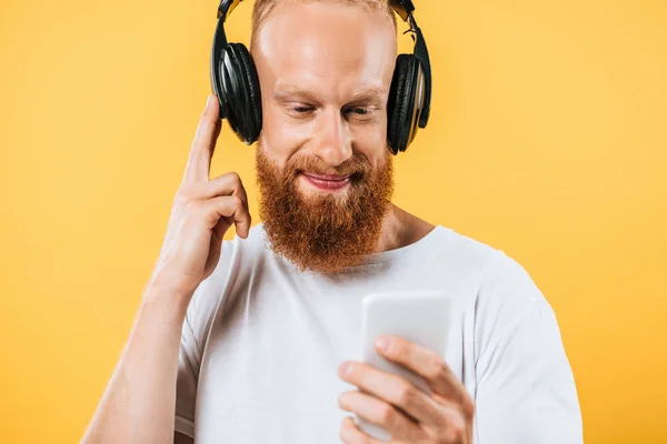 Uomo barba sorridente che ascolta musica con cuffie e smartphone, isolato su giallo — Foto stock