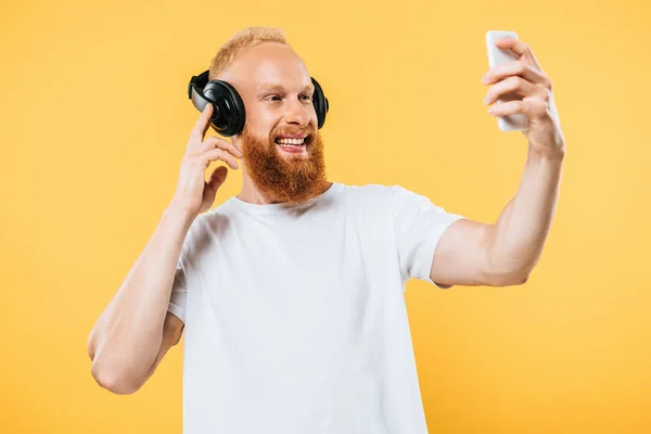 Barba hombre escuchar música con auriculares y tomar selfie en el teléfono inteligente, aislado en amarillo - foto de stock