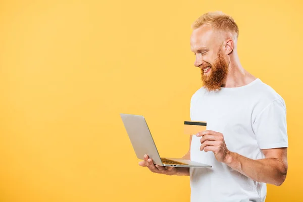 Веселый бородатый мужчина делает покупки в Интернете с ноутбуком и кредитной картой, изолированные на желтый — стоковое фото