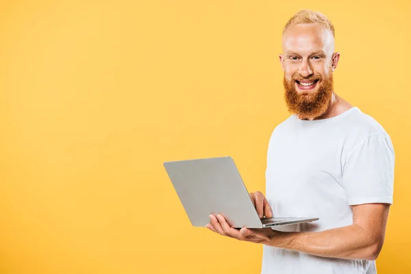 Homem barbudo sorridente usando laptop, isolado no amarelo — Fotografia de Stock