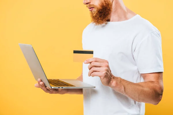 Vista recortada de hombre barbudo de compras en línea con ordenador portátil y tarjeta de crédito, aislado en amarillo - foto de stock