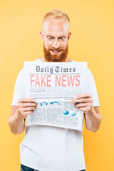 Homem barbudo sorridente lendo jornal com notícias falsas, isolado no amarelo — Fotografia de Stock