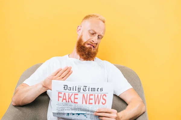 Бородатый мужчина с отвращением читает фейковые новости, сидя на кресле, изолированном на желтом — стоковое фото
