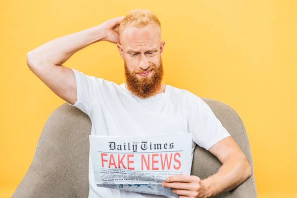 Вдумчивый бородатый мужчина читает газету с фальшивыми новостями, сидя на кресле, изолированном на желтом — стоковое фото