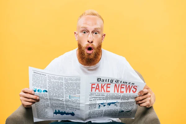 Homem surpreso lendo jornal com notícias falsas enquanto sentado em poltrona, isolado no amarelo — Fotografia de Stock