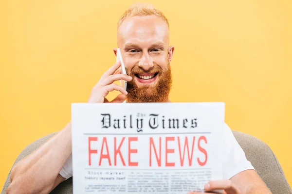 Alegre hombre leyendo el periódico con noticias falsas y hablando en el teléfono inteligente mientras está sentado en el sillón, aislado en amarillo - foto de stock