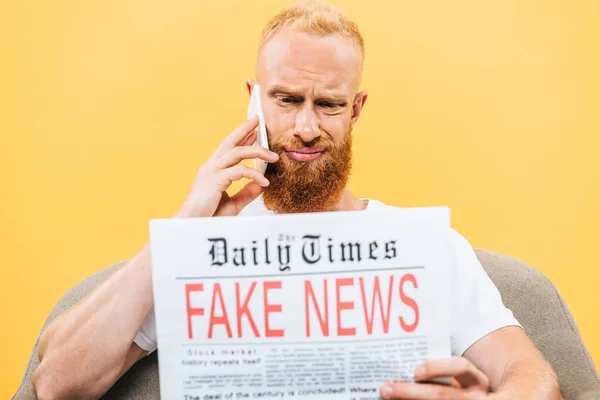 Escéptico hombre leyendo el periódico con noticias falsas y hablando en el teléfono inteligente mientras está sentado en el sillón, aislado en amarillo - foto de stock