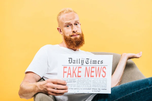 Скептик, читающий газету с фейковыми новостями, сидя на кресле, пожимая плечами, изолированный на желтом — стоковое фото