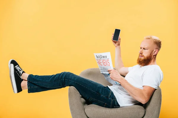 Homme sérieux lisant le journal avec de fausses nouvelles et montrant sur smartphone avec écran vide tout en étant assis sur un fauteuil, isolé sur jaune — Photo de stock