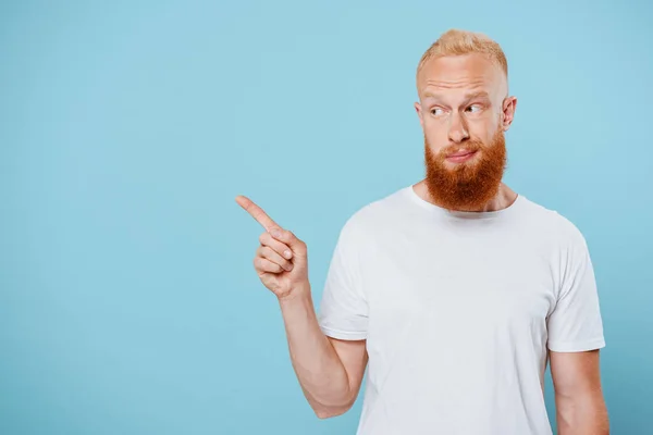 Ritratto di sarcastico uomo barbuto in t-shirt bianca che punta a qualcosa, isolato sul blu — Foto stock