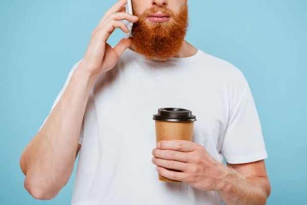 Vue recadrée de sérieux homme barbu avec du café pour aller parler sur smartphone, isolé sur bleu — Photo de stock