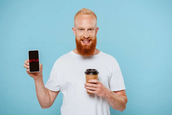 KYIV, UCRANIA - 27 de agosto de 2019: hombre barbudo sonriente con café para mostrar el teléfono inteligente con la aplicación netflix, aislado en azul - foto de stock