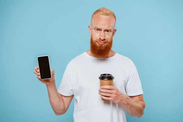 Homme barbu sceptique avec café pour aller montrant smartphone avec écran blanc, isolé sur bleu — Photo de stock