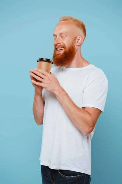 Hombre barbudo feliz en camiseta sosteniendo café para ir con los ojos cerrados, aislado en azul - foto de stock