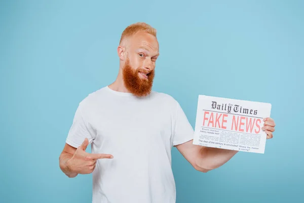 Sonriente barbudo apuntando al periódico con noticias falsas, aislado en azul - foto de stock