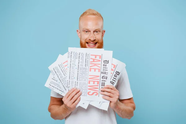 Happy bearded man holding newspapers with fake news, isolated on blue — Stock Photo