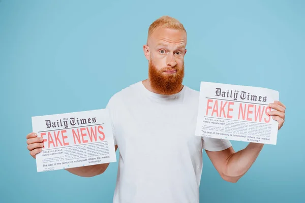 Hombre barbudo confundido sosteniendo dos periódicos con noticias falsas, aislado en azul - foto de stock