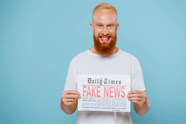 Hombre barbudo sonriente mostrando periódico con noticias falsas, aislado en azul - foto de stock