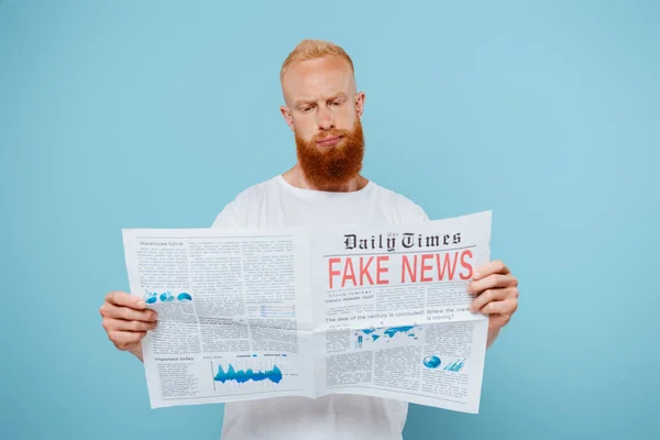 Hombre barbudo escéptico leyendo el periódico con noticias falsas, aislado en azul - foto de stock