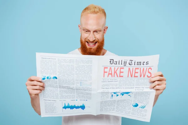 Hombre barbudo alegre leyendo el periódico con noticias falsas, aislado en azul - foto de stock